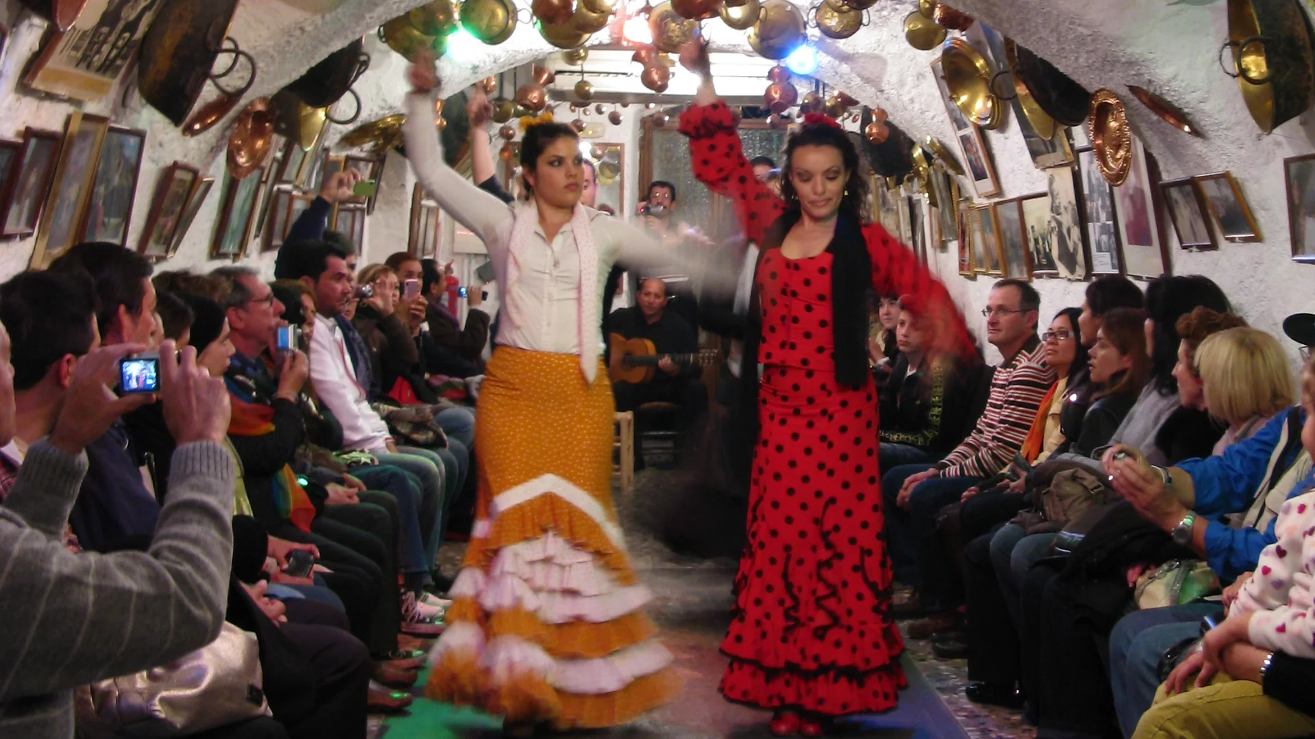 Dva tanečníci flamenca, jeden v bílé halence a okrové sukni, druhý v červených šatech s puntíky, vystupují ve vybílené jeskyni v Sacromonte ověšené měděnými hrnci, za nimi kytarista a hosté usedlí podél stěn