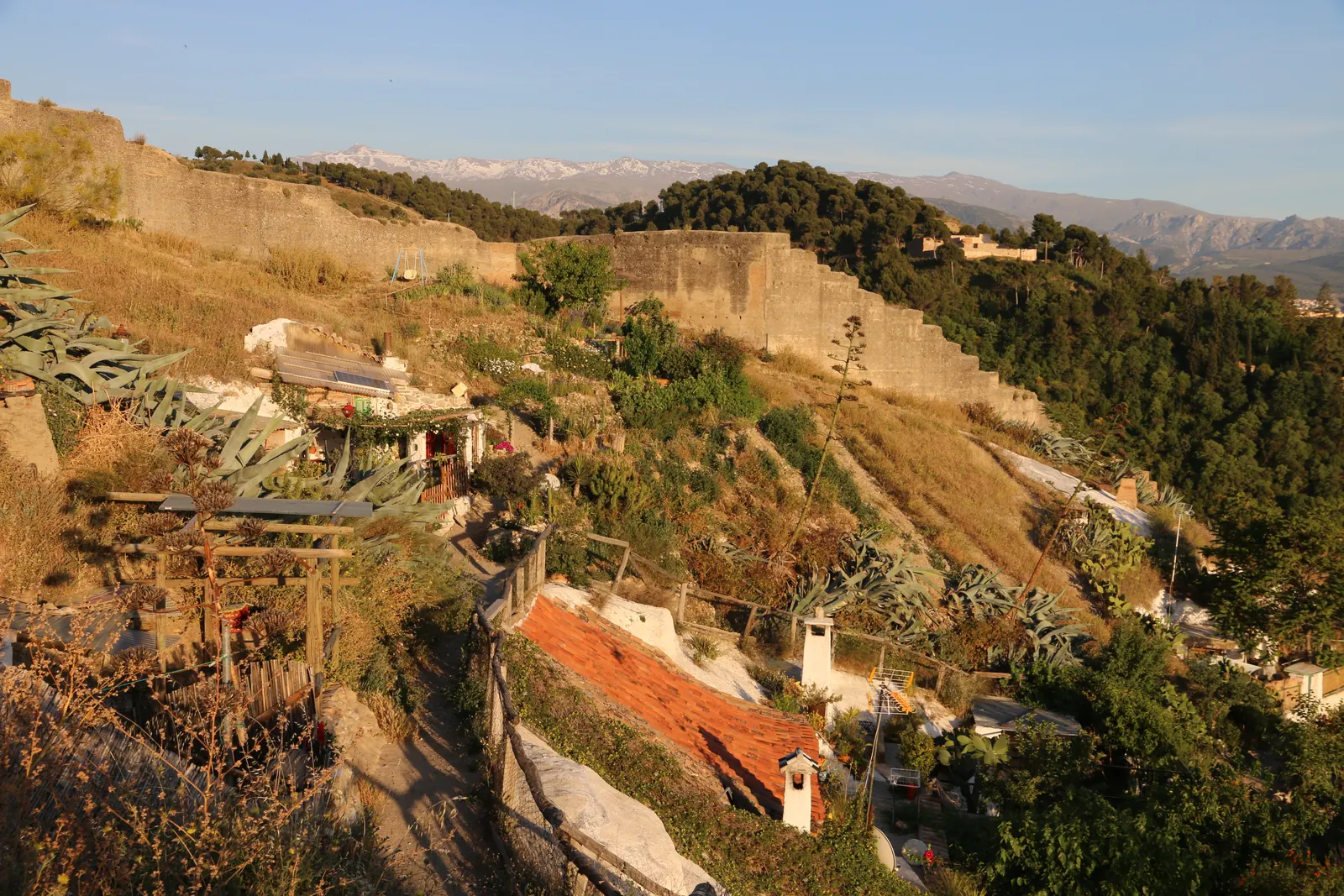 Jeskynní obydlí a zahrady na svahu Sacromonte, nad nimi kamenná zeď a v dálce Sierra Nevada