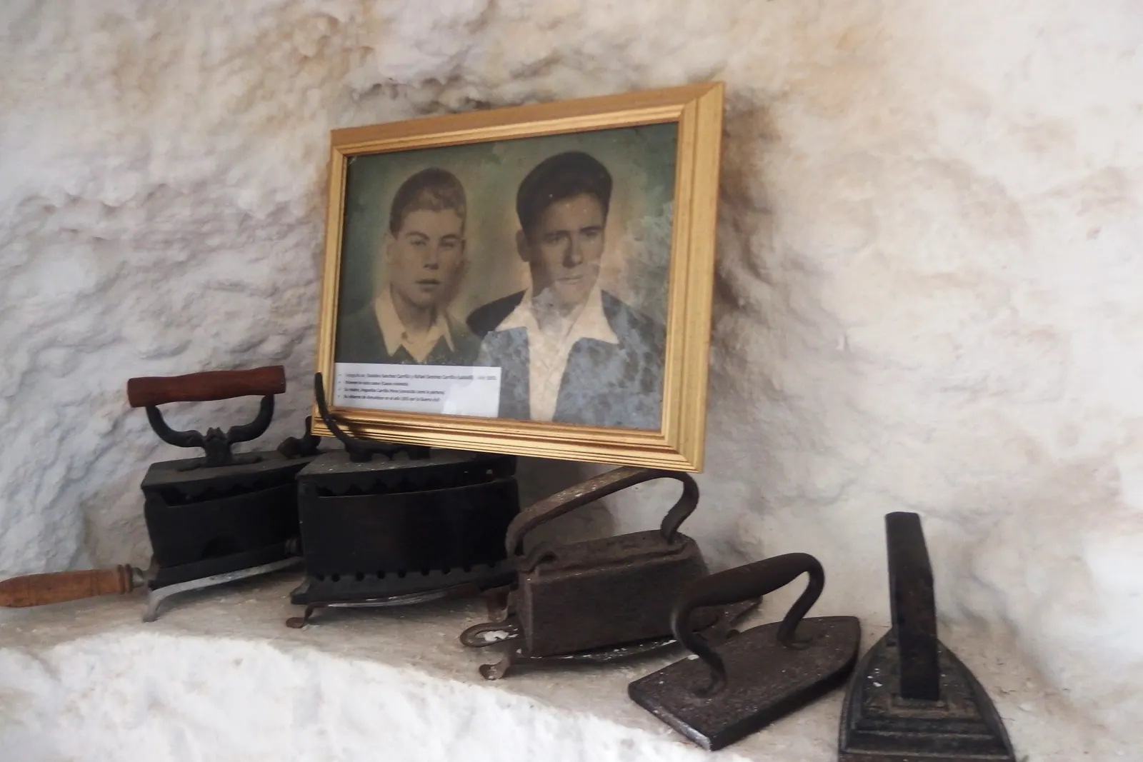 Staré žehličky a zarámovaná fotografie dvou mladých mužů na bíle obílené poličce v jeskyni v muzeu Sacromonte